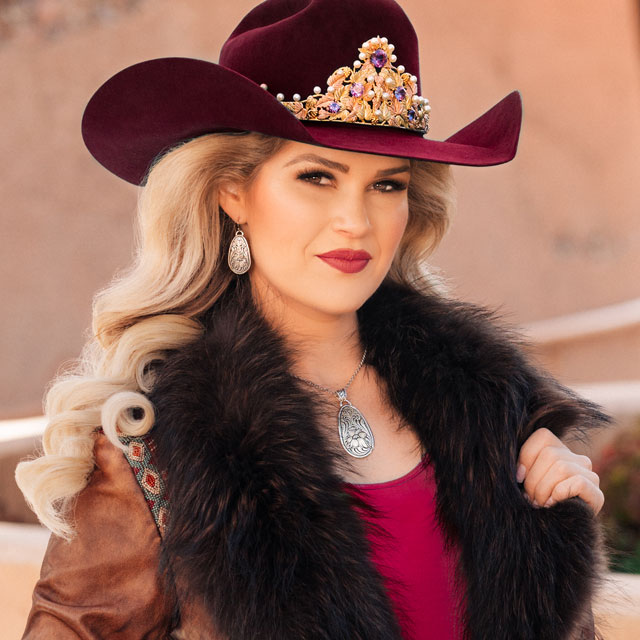 Miss Rodeo America is wearing a brown jacket with black fur around the neck/ collar and a maroon hat, posing for the camera.